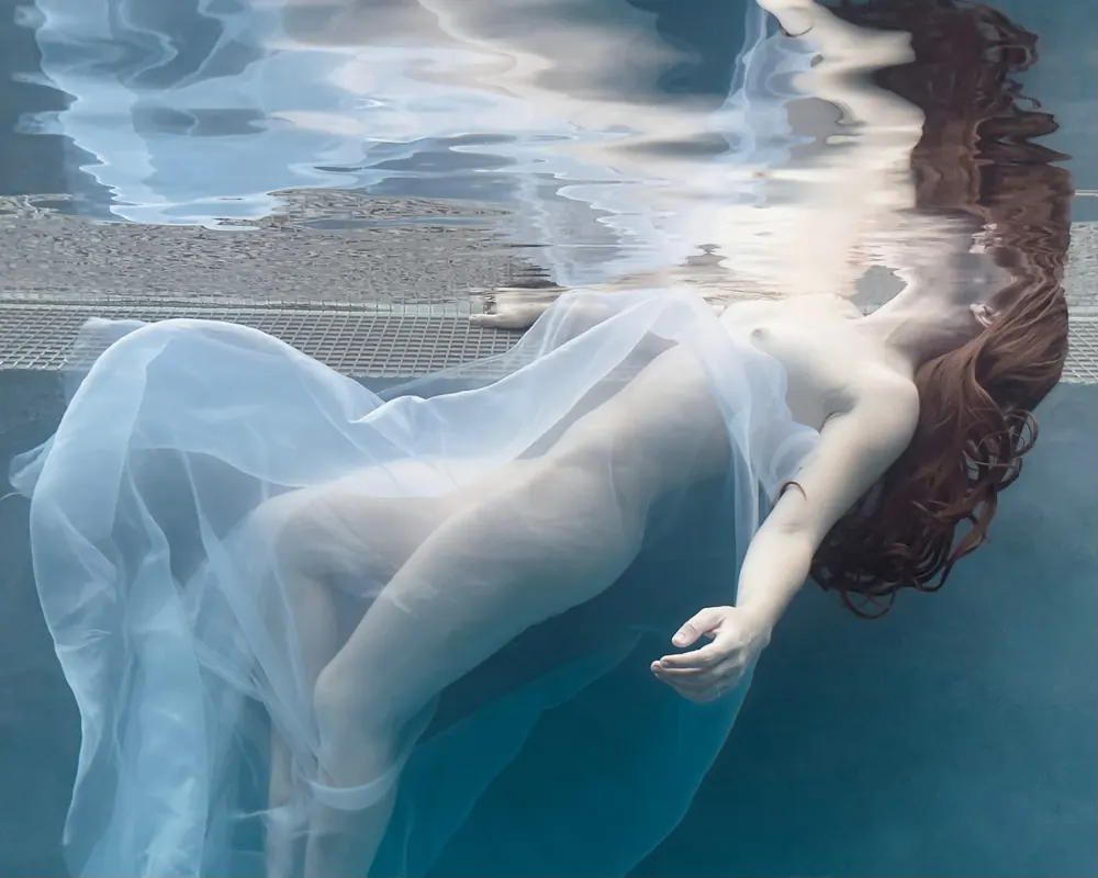 Surrender — a figure draped in sheer white fabric drifting beneath the surface of a pool